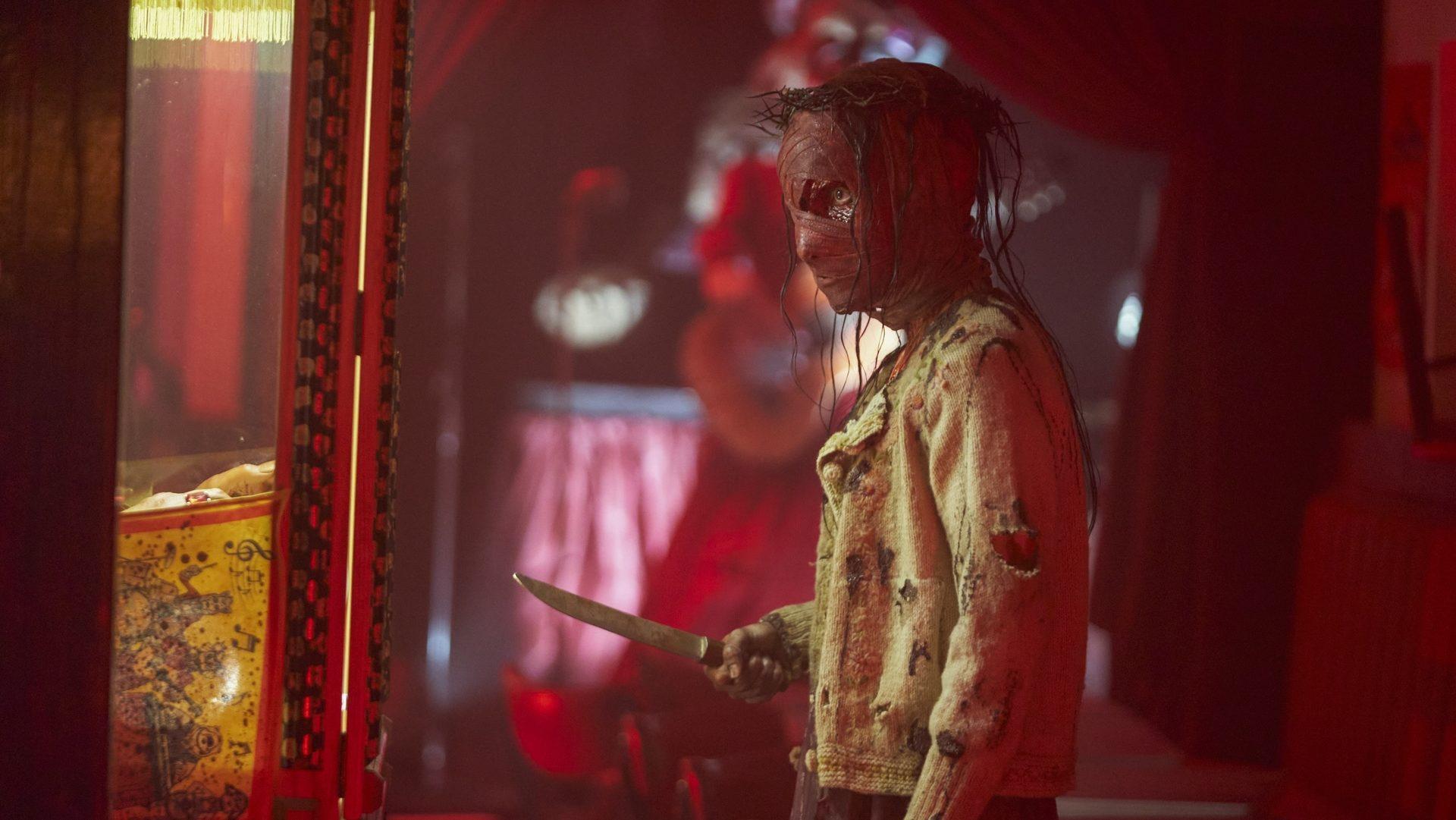A figure with red bandages covering their face holds a knife 