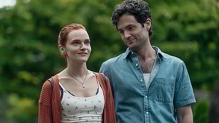 You A dark haired man stands next to a woman with red hair. He looks at her and smiles while she looks forward with a smile.