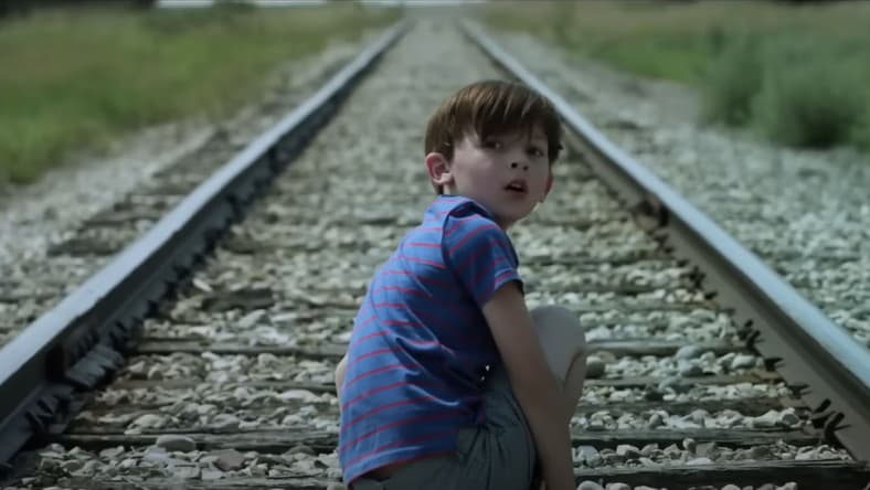 A screenshot from the film Z (2020) showing a young boy crouching down on railroad tracks looking over his shoulder at the camera. His face looks surprised.