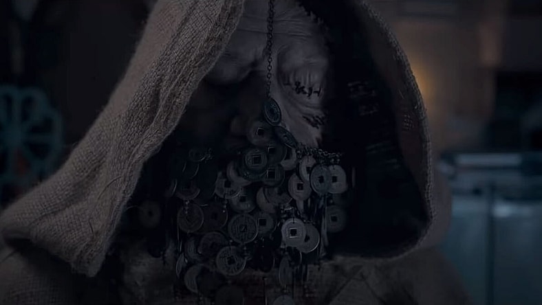 A roughly textured hood covers a shadowy figures head, their eyes have been stitched shut and a veil made of coins hangs over their mouth.