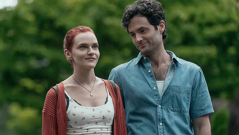 You A dark haired man stands next to a woman with red hair. He looks at her and smiles while she looks forward with a smile.