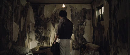 A screenshot from the film 'The Chaser' of a man standing in front of a wall with peeling wallpaper.