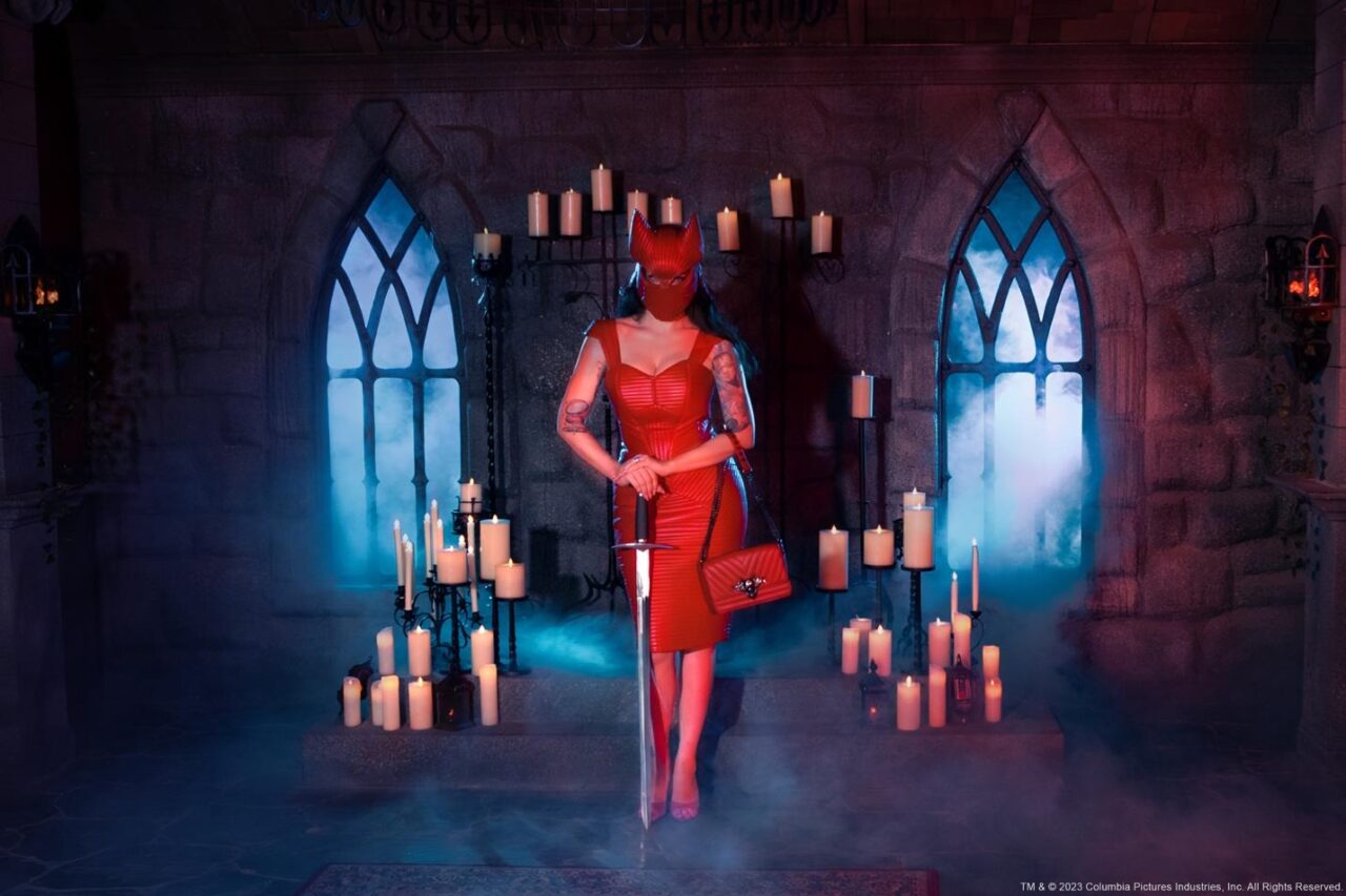 A person in a red dress holding a sword in front of a stone building with candles

Description automatically generated