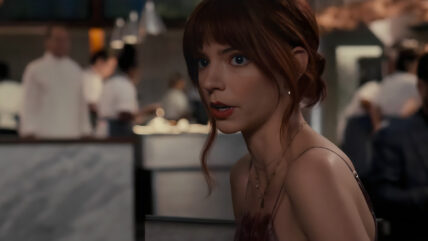 Anna Taylor-Joy from the film The Menu. She sits at a table with her often rude date while the chefs can be seen in the background behind her preparing the twisted meals of The Menu.