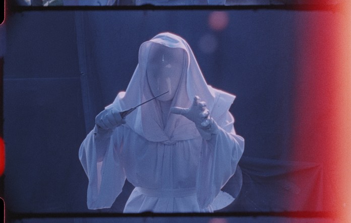 A figure in a white robe, white hood, and white mask that completely obscures their face holds a knife menacingly in one hand and reaches forward with the other.