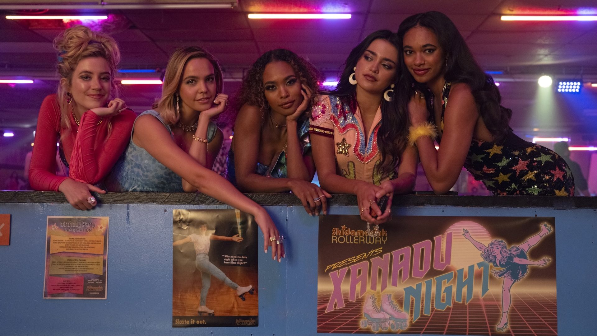 A group of five teenage girls lean on the side of a roller rink while wearing 80s style outfits 